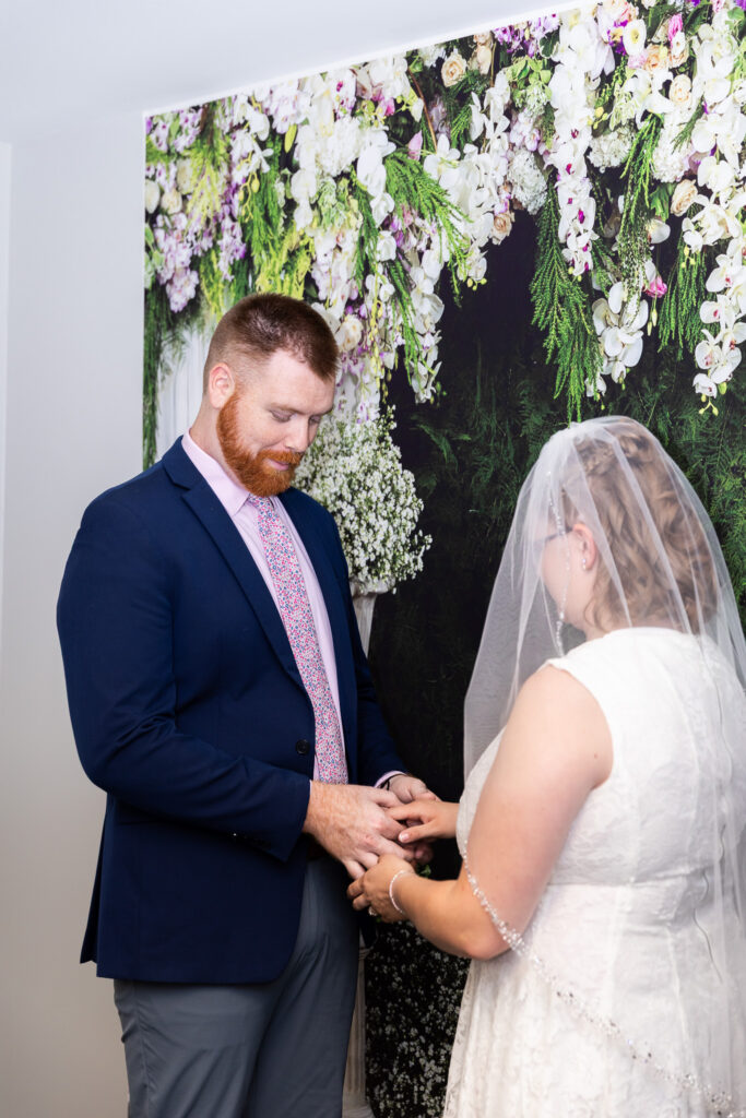 Bride in a short white lace dress and veil kisses the groom’s cheek during courthouse wedding photography, framed by a lush backdrop of white flowers and greenery.