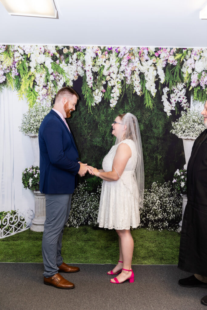Couple faces each other and holds hands during their intimate civil ceremony, surrounded by white and lavender flowers, greenery and small arrangements of baby’s breath.