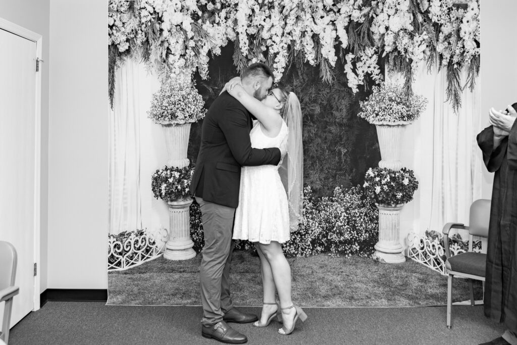 A black and white capture of a newly married couple sharing their first kiss during a Pennsylvania civil ceremony, an emotional moment captured through courthouse wedding photography in front of a floral and greenery backdrop.