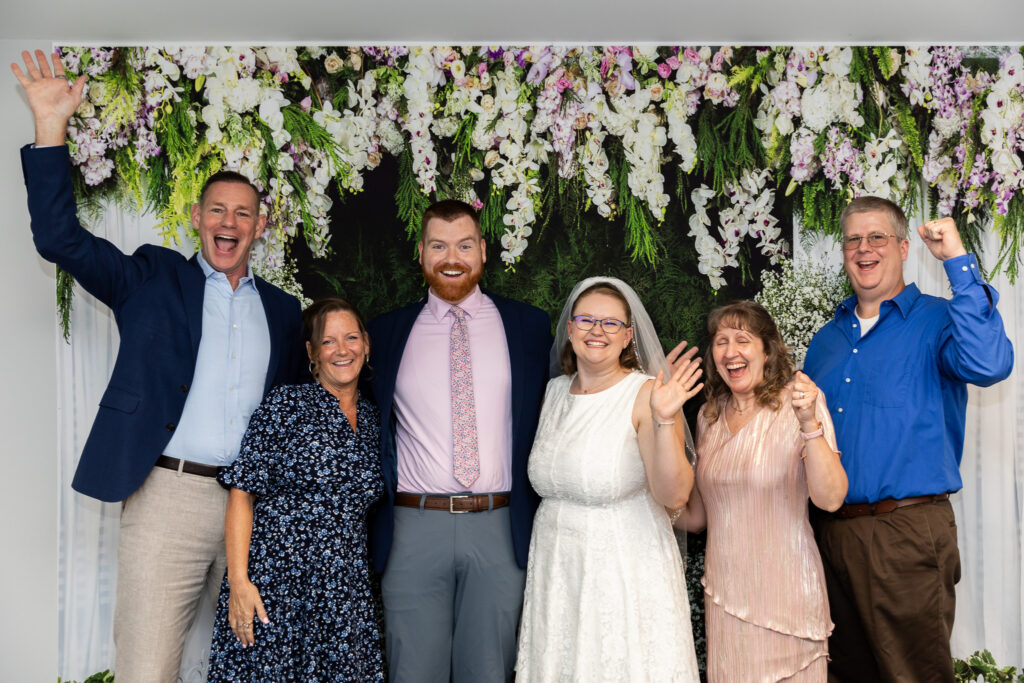 Newlyweds celebrate their courthouse wedding with their parents, everyone smiling, cheering and raising their hands together in front of an elaborate floral backdrop.