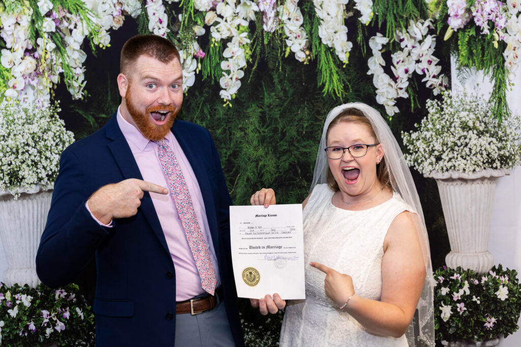 Newly married couple excitedly points to their signed marriage license in a courthouse wedding photo, standing beneath hanging white flowers and lush greenery.