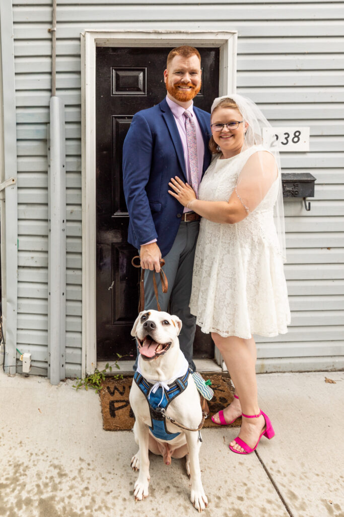 Newlyweds pose outside with their white dog after their intimate courthouse elopement, the bride wearing a short lace dress, veil and bright pink heels beside the groom in a navy jacket.
