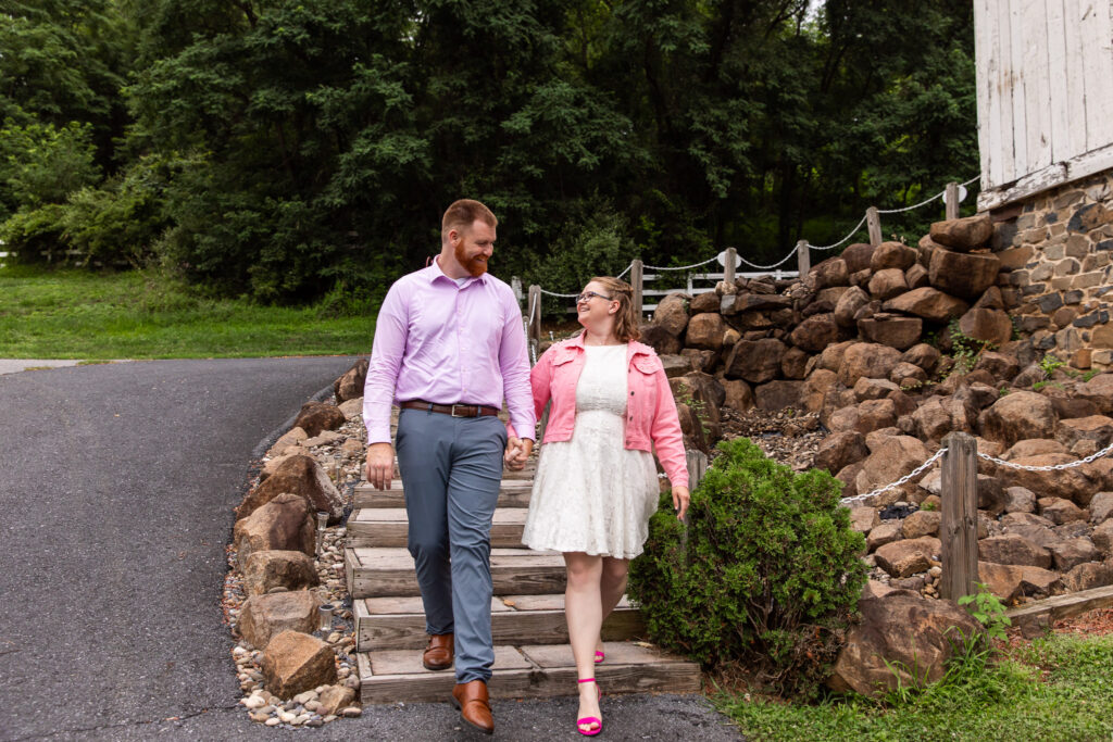 Couple walks hand in hand down wooden steps during their engagement photos at Pleasant View Farm, smiling at each other beside a rustic stone wall and wooded landscape.