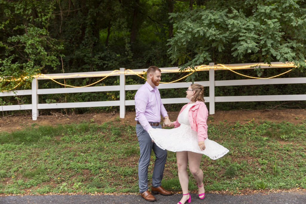 Couple laughs together during playful couples portraits at Pleasant View Farm as the bride twirls her white lace dress beside a fence lined with string lights.