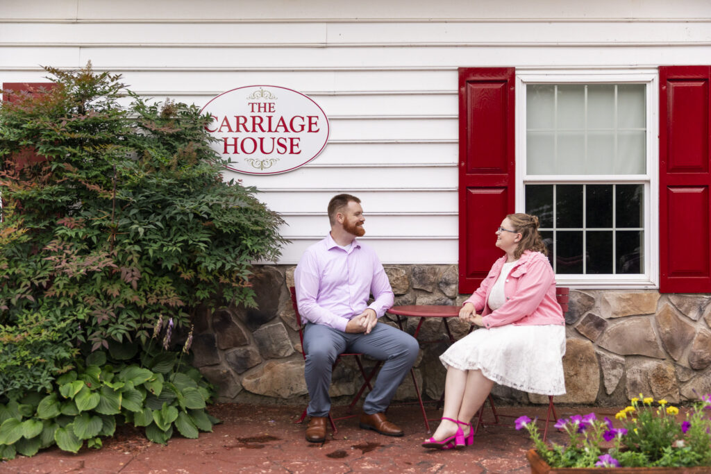 Couple sits facing each other beneath The Carriage House sign during their engagement session, surrounded by red shutters, stonework, flowers and lush landscaping.