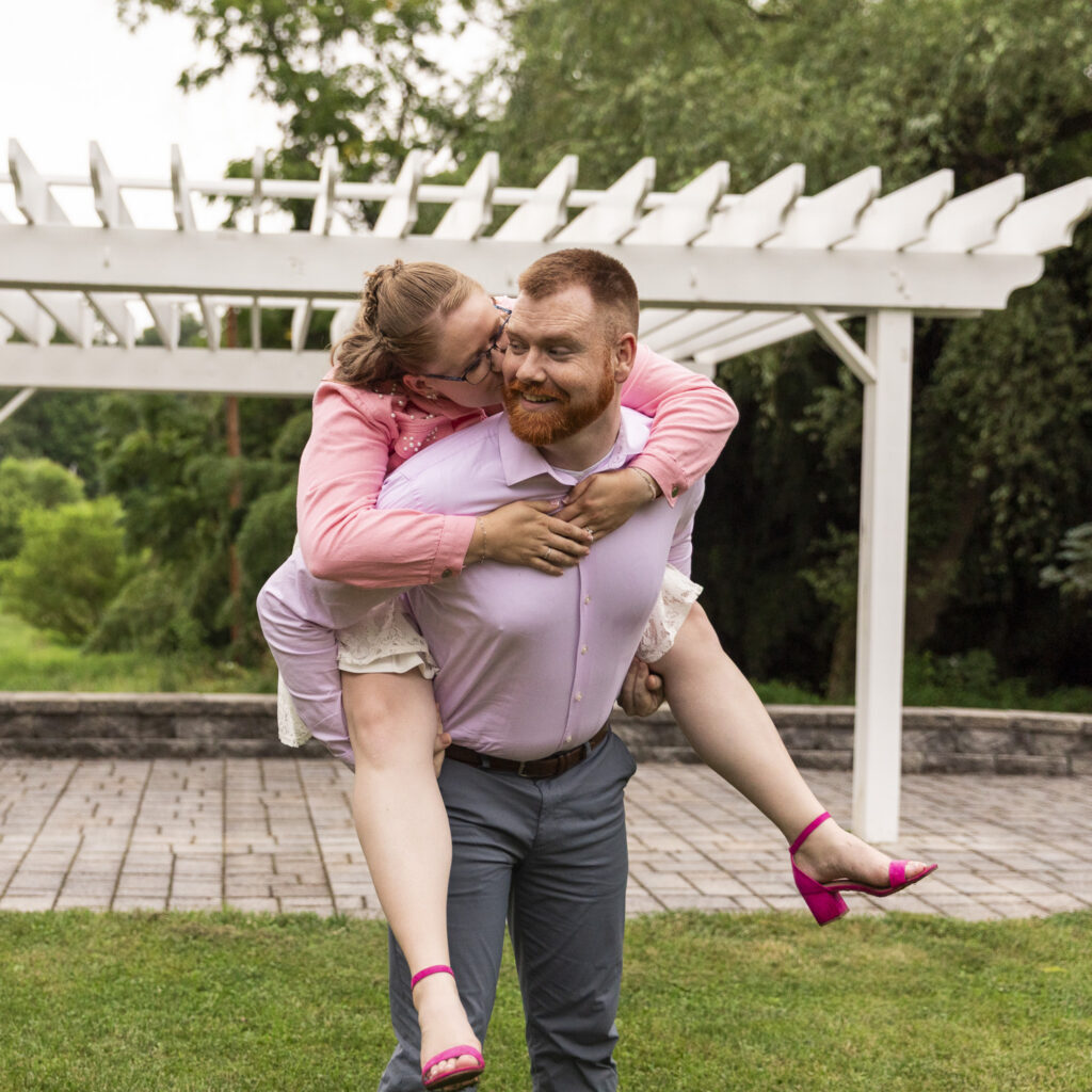 A bride rides piggyback and kisses the groom’s cheek during playful engagement photos beneath a white pergola at Pleasant View Farm.
