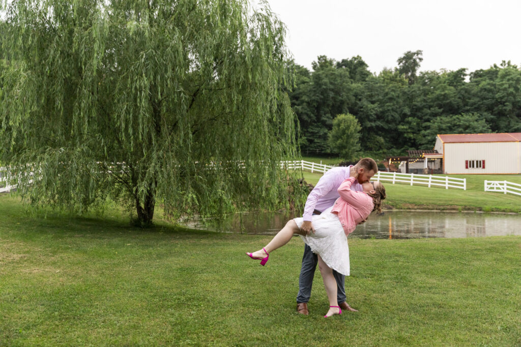 Groom dips and kisses the bride during their engagement session beside a pond and large willow tree at Pleasant View Farm, with white fencing and the venue grounds in the distance.