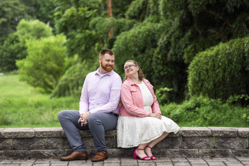 Couple sits side by side on a low stone wall during their engagement photos, smiling together with a lush green field and trees stretching into the background.