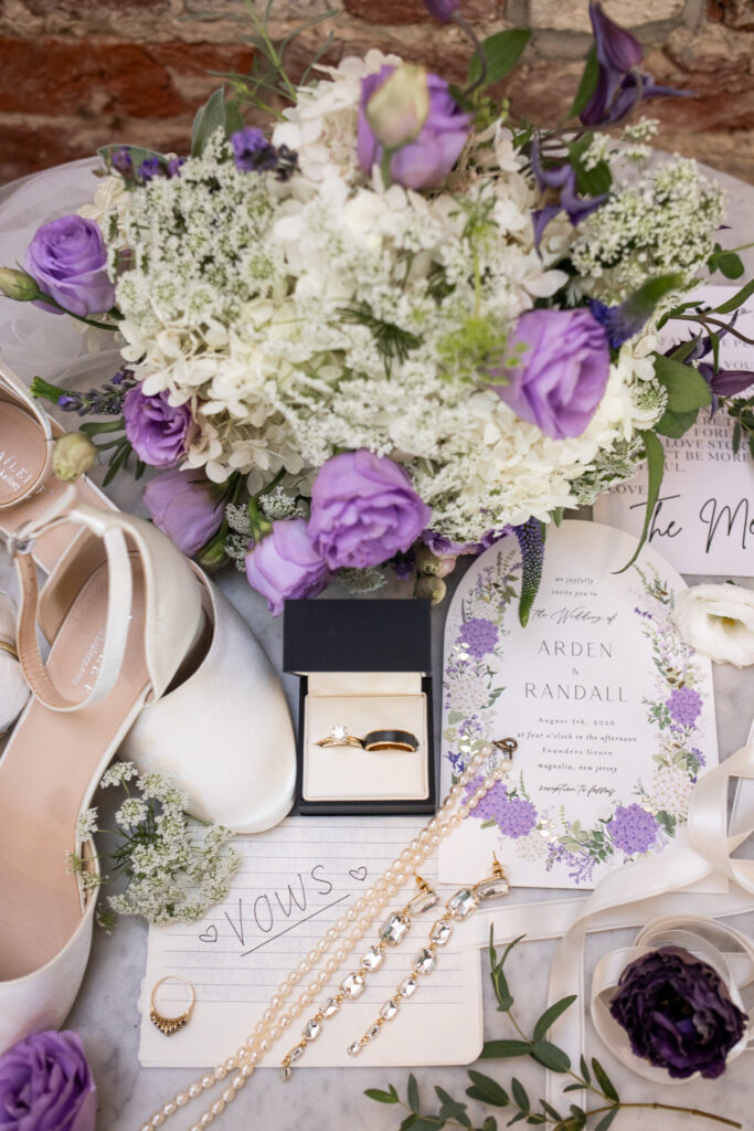 A flat lay of wedding details features a white and purple bouquet, invitation suite, rings, pearl necklace, jewelry and ivory heels arranged together for a South Jersey wedding.