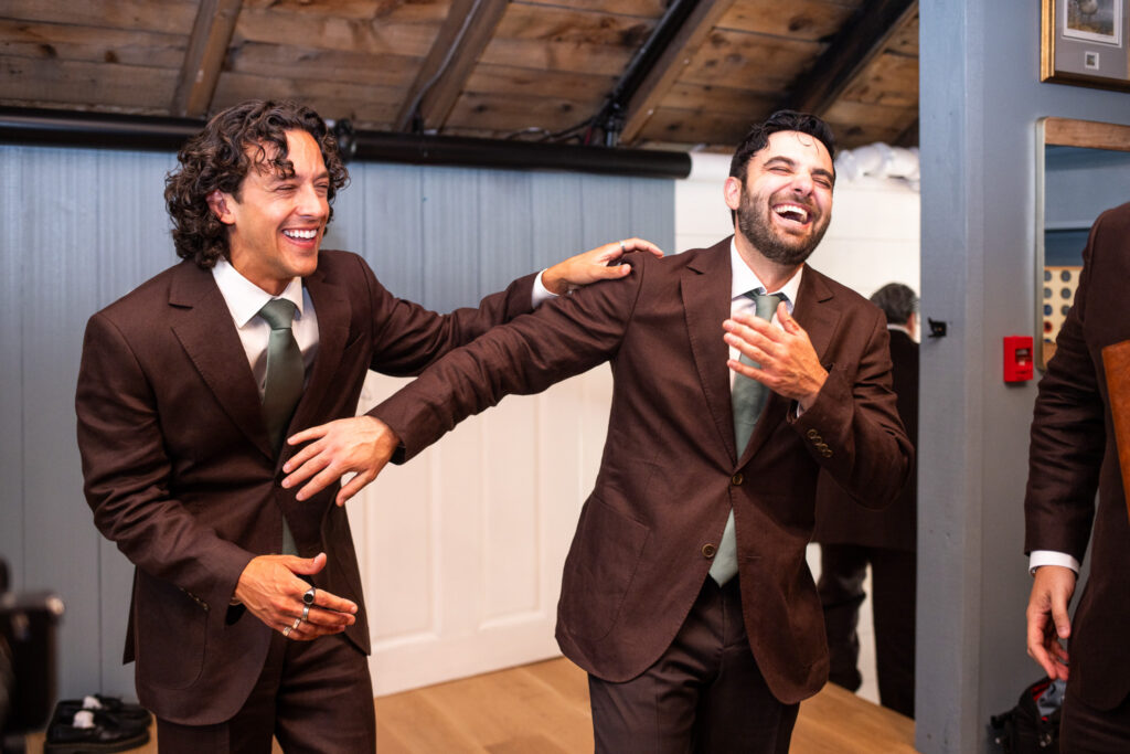 The groom and a groomsman in dark brown suits and sage green ties laugh together while getting ready inside a Magnolia, NJ wedding venue, one reaching toward the other as they share the moment.