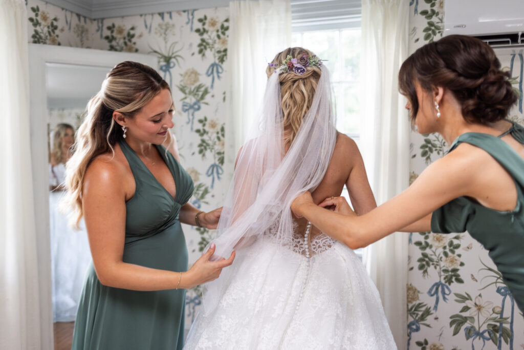 Two bridesmaids in sage green dresses help fasten the back of the bride’s lace ball gown and arrange her fingertip-length veil inside a bright floral-wallpapered room at a Magnolia, NJ wedding venue.