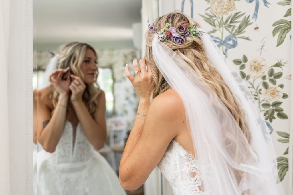 A bride adjusts her earring in a mirror during her Founders Grove wedding, wearing a floral hairpiece and fingertip veil against the farmhouse’s botanical wallpaper.