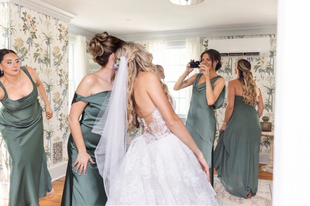A bride poses for a photo taken by a bridesmaid with her wedding party in sage green dresses inside a bright room with botanical wallpaper during a South Jersey wedding.
