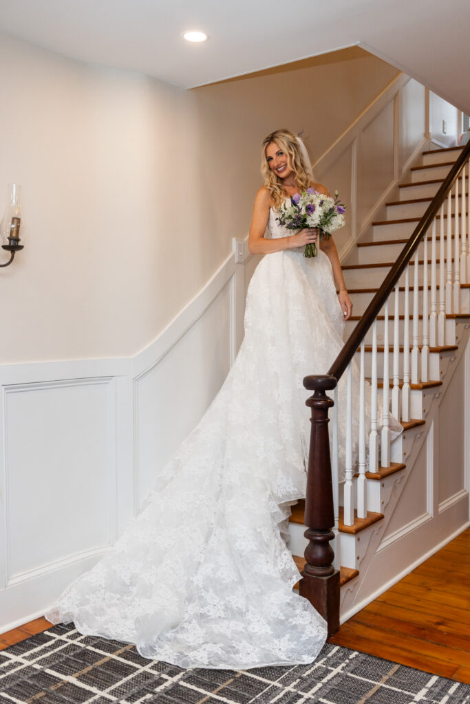 A bride in a strapless lace wedding gown holds a purple and white bouquet while posing on the wooden staircase of a historic farmhouse at a South Jersey wedding.