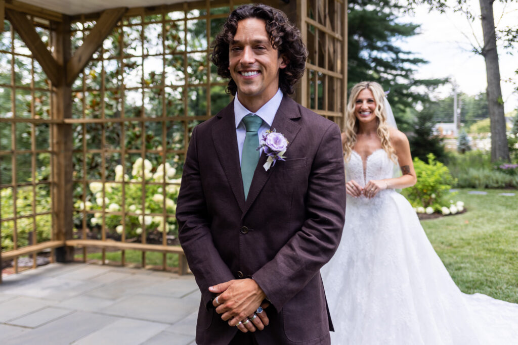 A groom in a dark brown suit and sage green tie smiles in the foreground while the bride approaches from behind during their outdoor first look at a Magnolia, NJ wedding venue, surrounded by gardens and a wooden structure.
