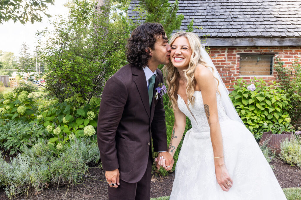 A groom kisses his smiling bride on the cheek during their Founders Grove wedding, surrounded by lush garden greenery beside a historic brick outbuilding.