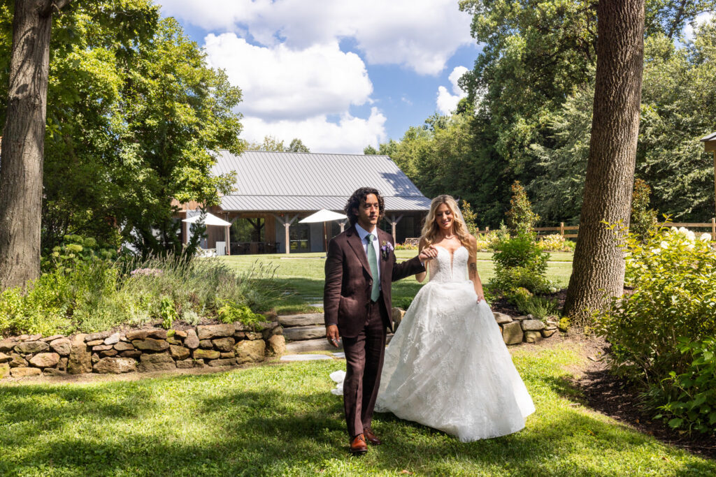 A newlywed couple walks through the landscaped grounds during their Founders Grove wedding, with the Carriage House, mature trees, and gardens visible behind them.