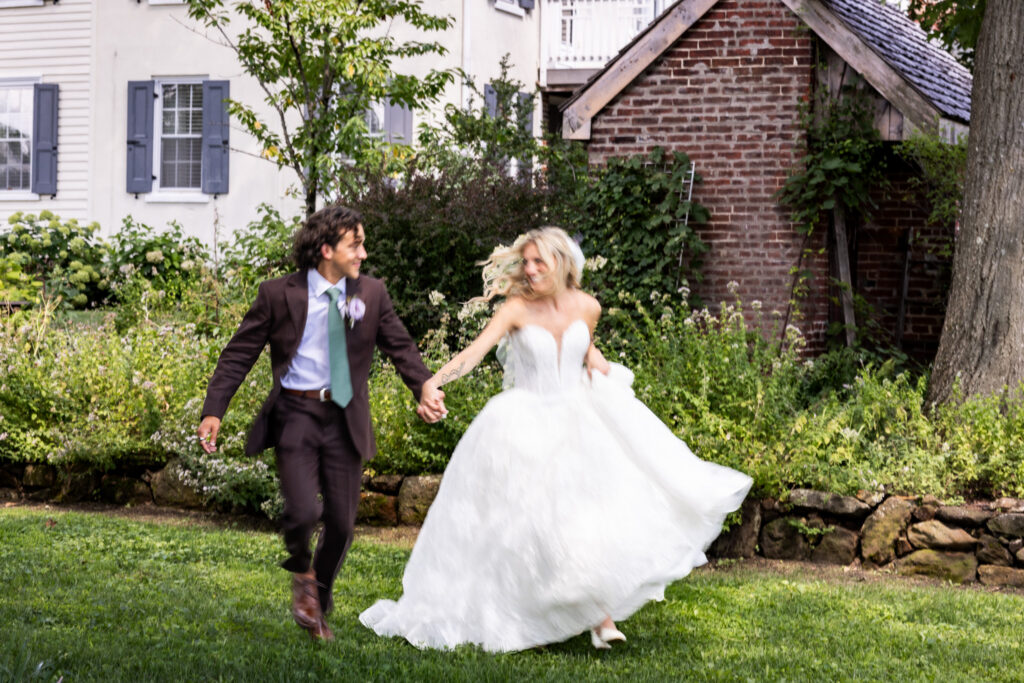 A blurry photo of a newlywed couple holds hands and runs across the lawn of a Magnolia, NJ wedding venue, the bride lifting her full lace skirt as they move past lush gardens, a historic brick outbuilding and farmhouse.