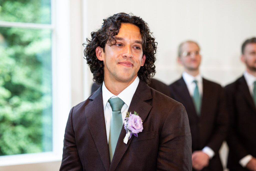 A groom wearing a dark brown suit, sage green tie and purple boutonniere becomes emotional during the indoor wedding ceremony as members of his wedding party stand behind him.
