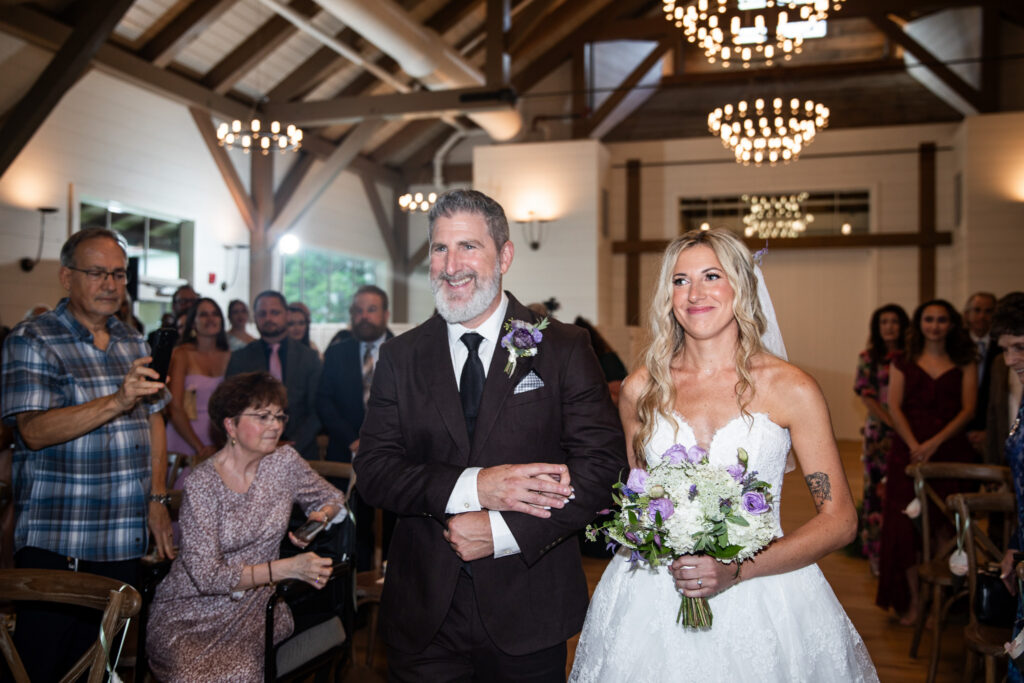 A bride walks down the aisle with her father during an indoor Founders Grove wedding ceremony, surrounded by guests beneath exposed wooden beams and glowing chandeliers.