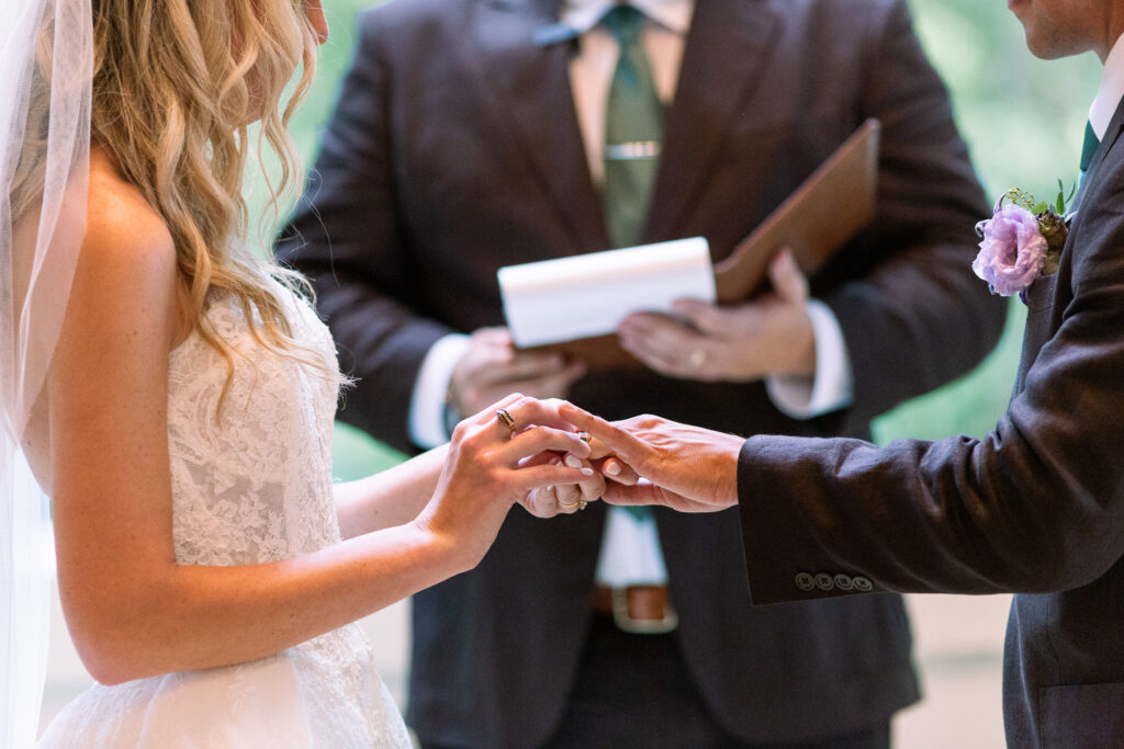 Close-up of a bride placing a wedding band onto the groom’s finger during their ceremony at a Magnolia, NJ wedding venue, with the officiant softly out of focus behind their hands.