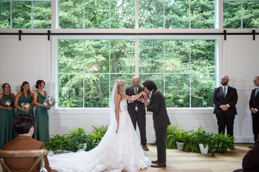 A groom kisses his bride’s hand during their Founders Grove wedding ceremony, framed by a wall of oversized windows overlooking the wooded property.