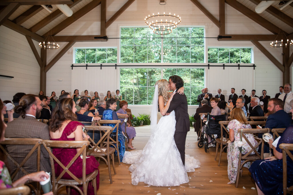 Newlyweds kiss in the center aisle after their Founders Grove wedding ceremony as seated guests cheer beneath the Carriage House’s vaulted beams and chandeliers.