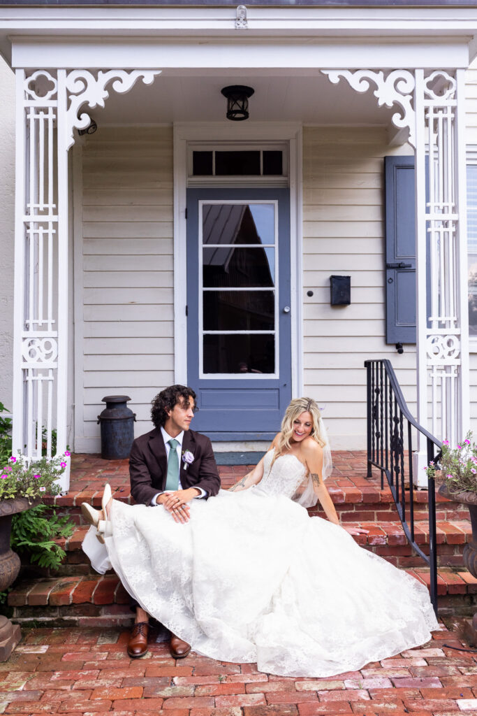 Newlyweds sit together on the brick steps of the historic farmhouse during their Founders Grove wedding, framed by ornate white porch trim and gray shutters.