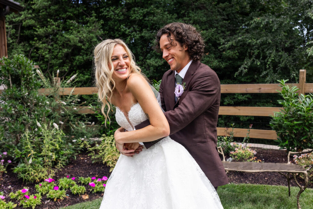 A bride and groom laugh together as he wraps his arms around her from behind during relaxed couple portraits in a lush garden.
