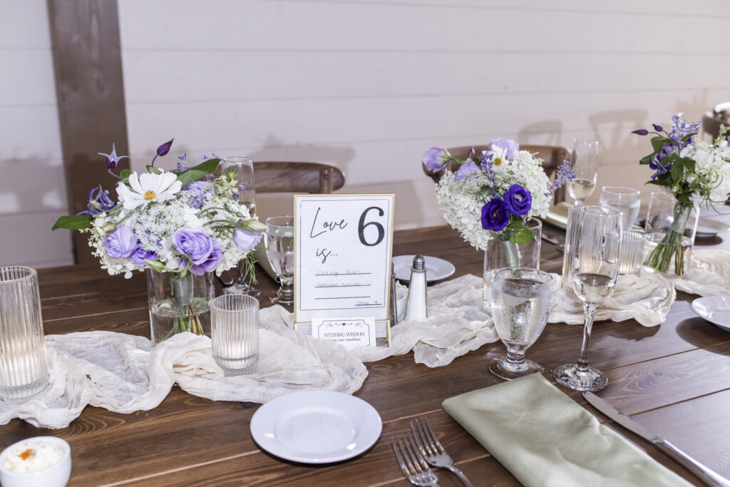 Purple and white floral centerpieces decorate a wooden reception table at a Founders Grove wedding, paired with sage napkins, glassware, candles, and a handwritten table sign.