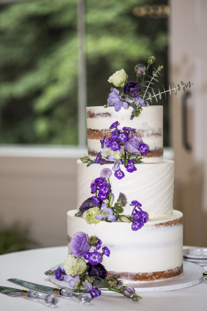 A three-tier semi-naked wedding cake decorated with fresh purple, lavender and white flowers sits beside crystal-handled cake servers at a South Jersey wedding.