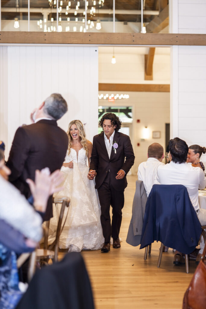 Newlyweds hold hands and smile as they enter their reception together between tables of applauding wedding guests.
