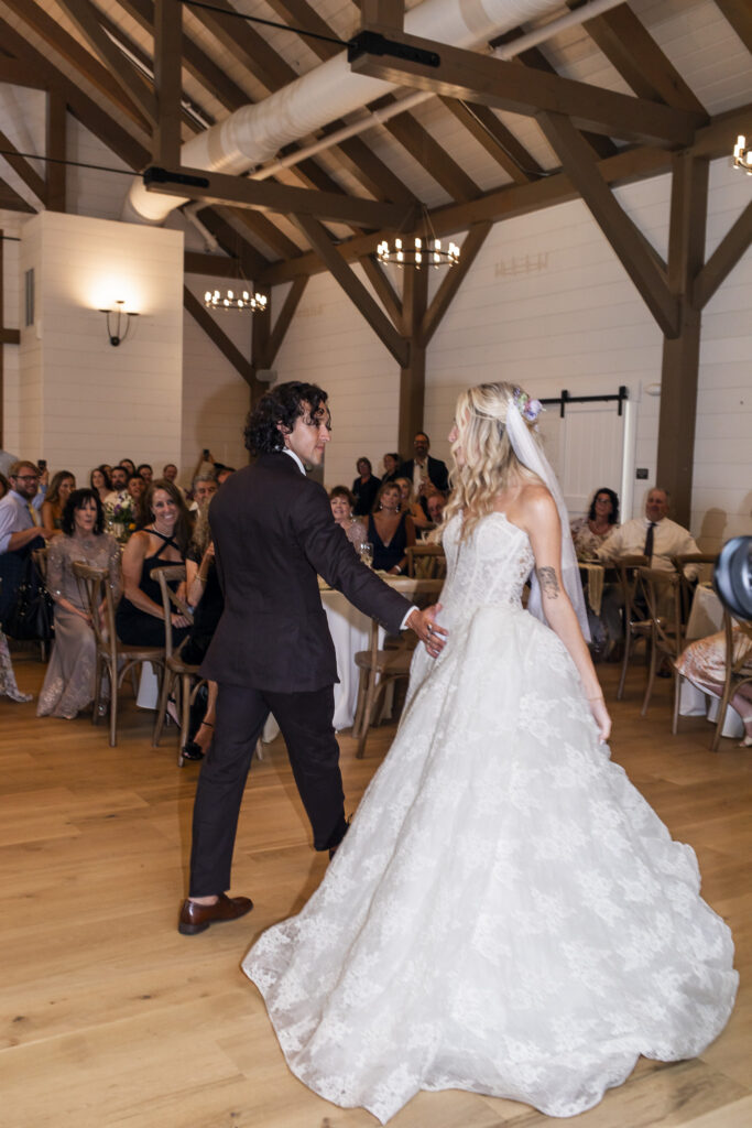 Newlyweds begin their first dance at their Founders Grove wedding reception hand in hand inside the Carriage House, with guests seated around them beneath exposed timber beams.