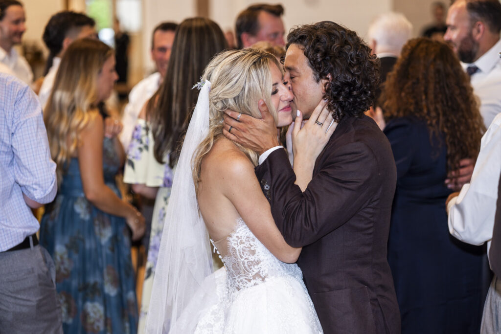A newlywed couple kisses on a crowded dance floor surrounded by guests during the reception at their South Jersey wedding.