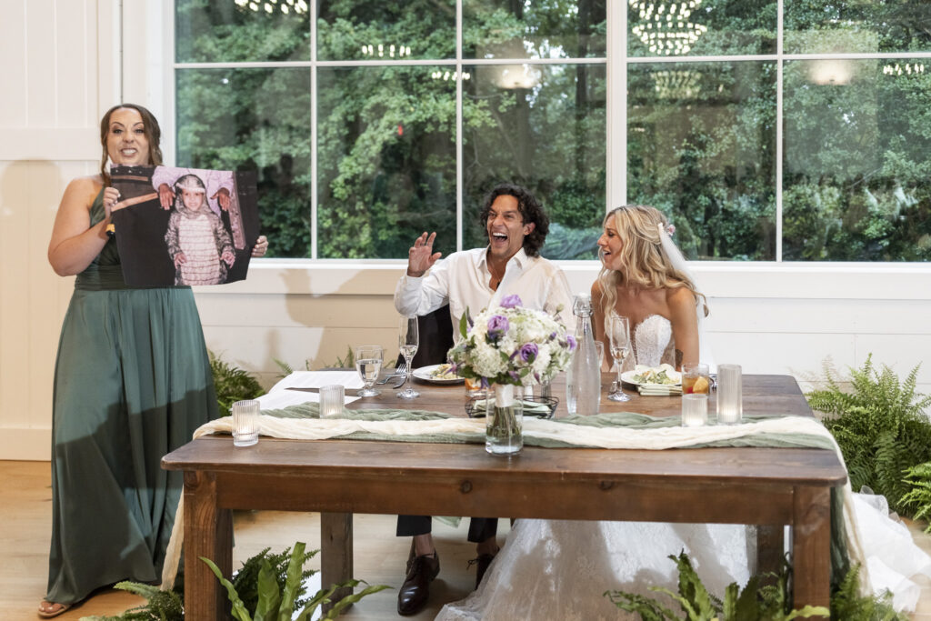 A wedding guest gives a humorous speech during a Founders Grove wedding reception, holding up a childhood photograph as the newlyweds laugh at their sweetheart table.