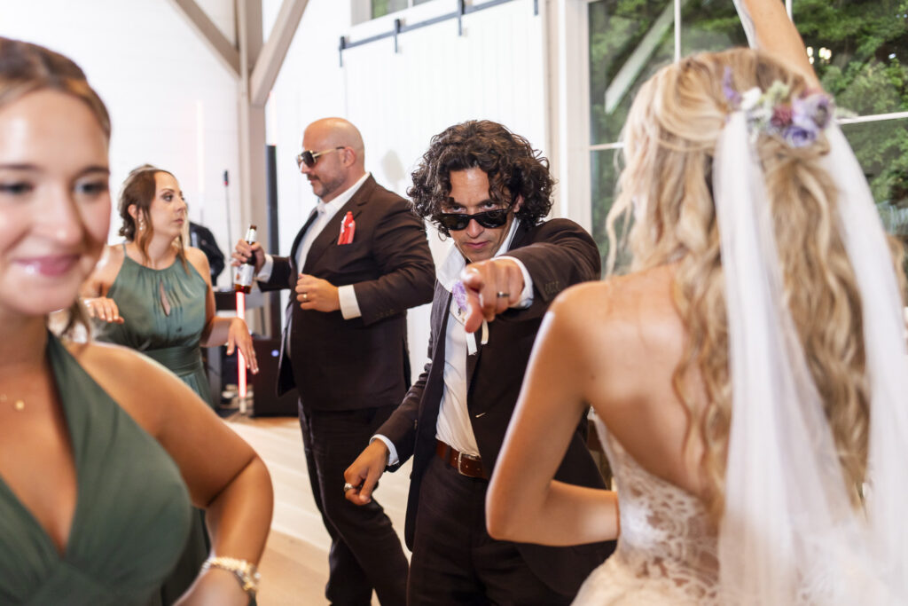 A groom in sunglasses points at the camera while dancing with friends inside the Carriage House during a lively South Jersey wedding reception.