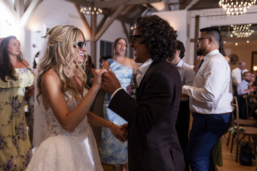 Newlyweds dance together during their Founders Grove wedding reception, wearing sunglasses while friends and family gather around them on the dance floor.