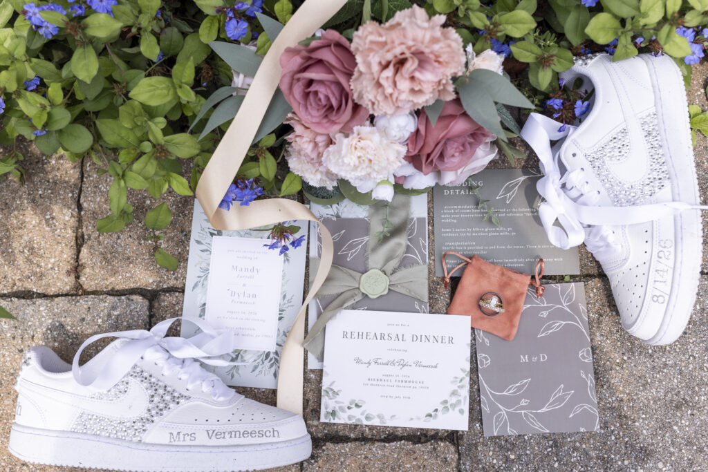 Pink and mauve wedding bouquet, invitation suite, rings and customized white sneakers arranged among blue flowers for a Tyler Arboretum wedding.