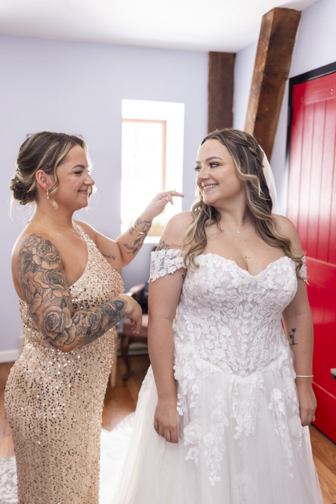 A bridesperson buttons the back of an off-the-shoulder lace wedding gown as the bride smiles beside a bright red door while getting ready.