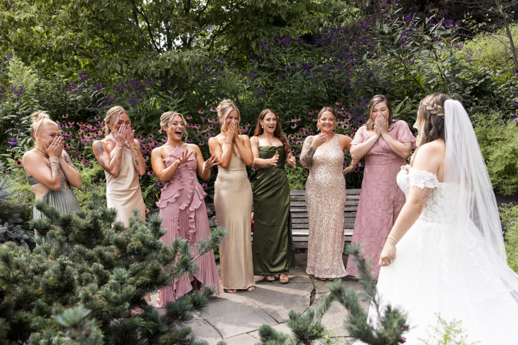 Bride reveals her wedding dress to a group of friends gathered in a lush garden filled with pink and purple flowers at Tyler Arboretum.