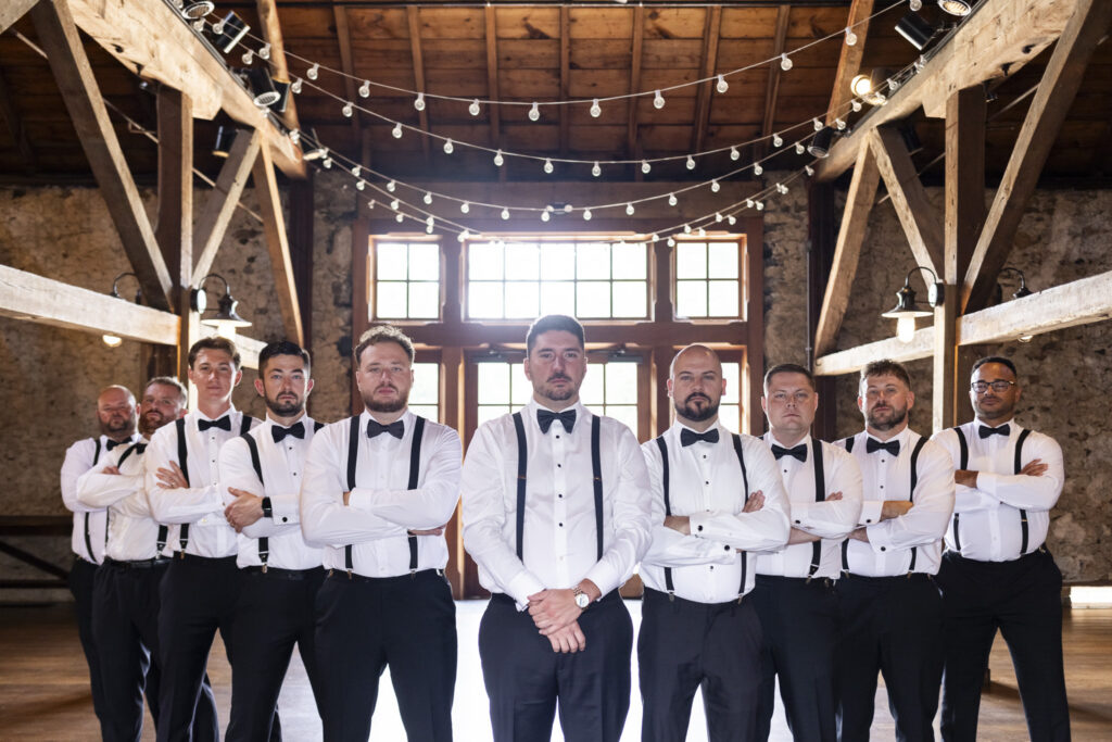 Groom and his wedding party pose inside a rustic stone barn with exposed wooden beams and string lights during a Tyler Arboretum wedding.