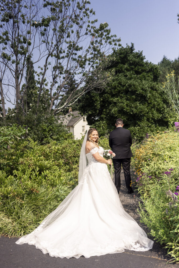 Bride approaches the groom from behind for their first look along a narrow garden path surrounded by colorful summer flowers at Tyler Arboretum.