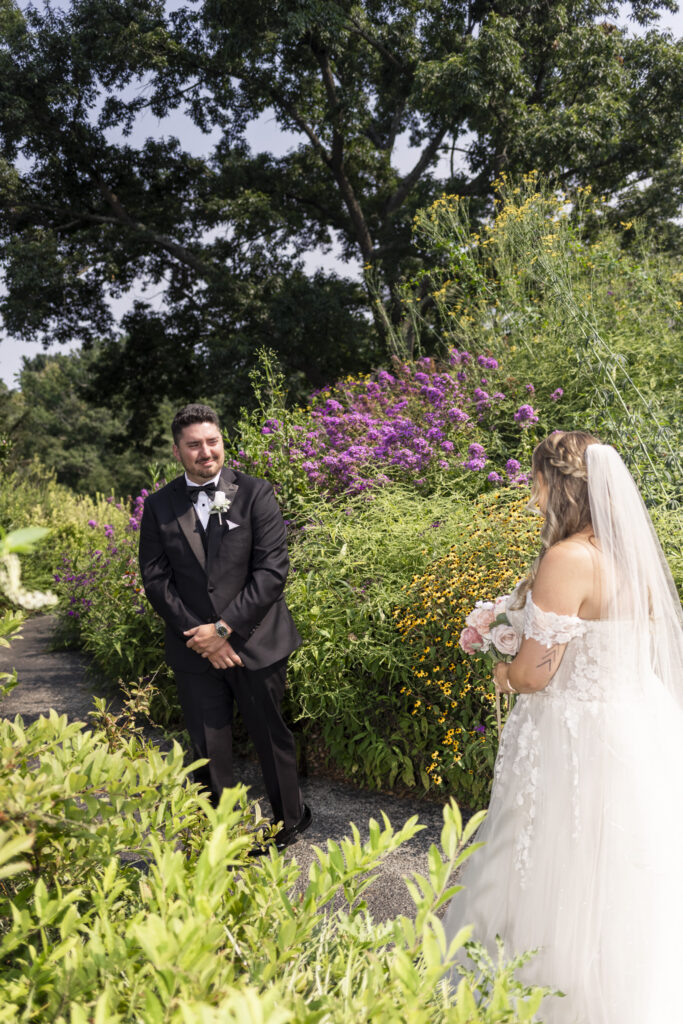 Groom smiles at the bride during their first look on a garden path surrounded by purple, yellow and green flowering plants.