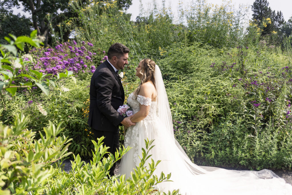 The newlyweds walk hand in hand along a narrow garden path surrounded by tall greenery and purple flowers, smiling at one another while the groom carries a pink and white bouquet during their Pennsylvania garden wedding.