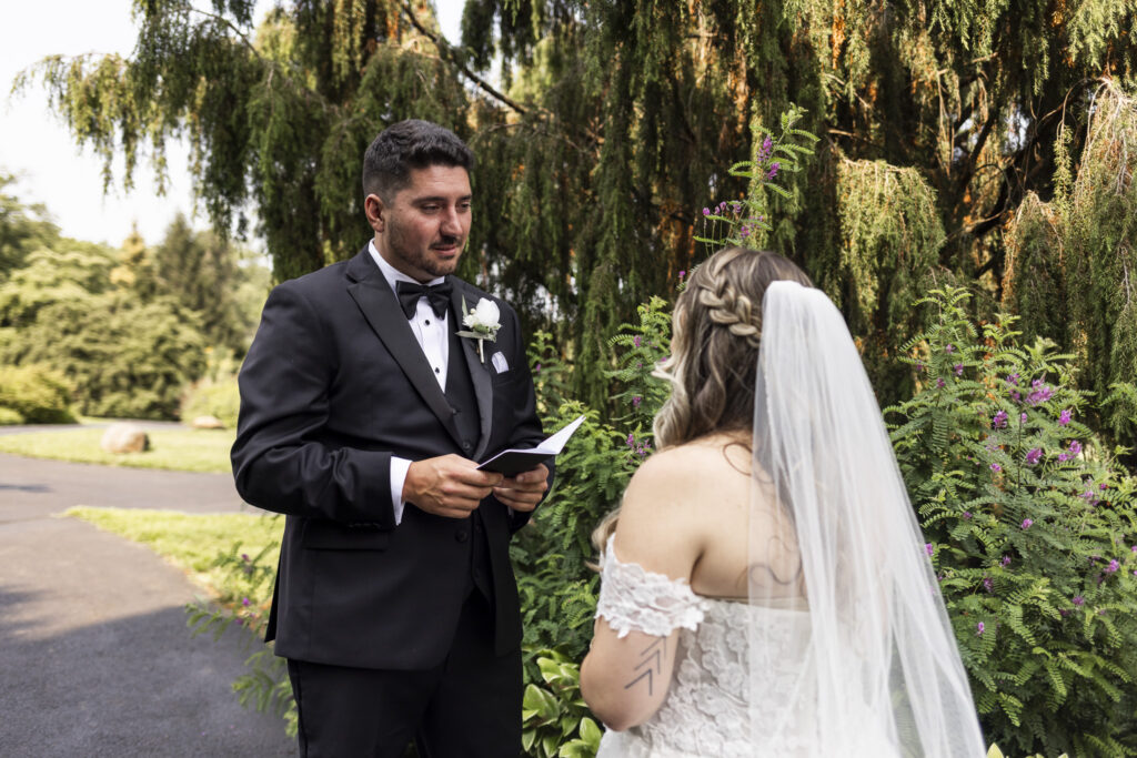 The groom reads from a small vow book while facing the bride during a private wedding-day moment in the gardens. She stands with her back toward the camera, showing her braided hairstyle, long veil, lace wedding dress, and arm tattoos while flowering greenery surrounds them.