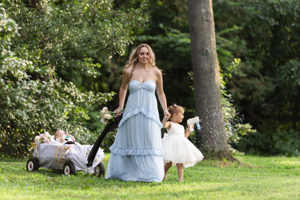 A member of the wedding party in a pale blue floor-length dress walks across a sunlit lawn, pulling an infant in a small decorated wagon while holding hands with a toddler in a white dress blowing bubbles during a Pennsylvania garden wedding.