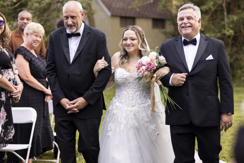 Bride walks down the outdoor ceremony aisle between two family members while carrying a pink and white bouquet at Tyler Arboretum.