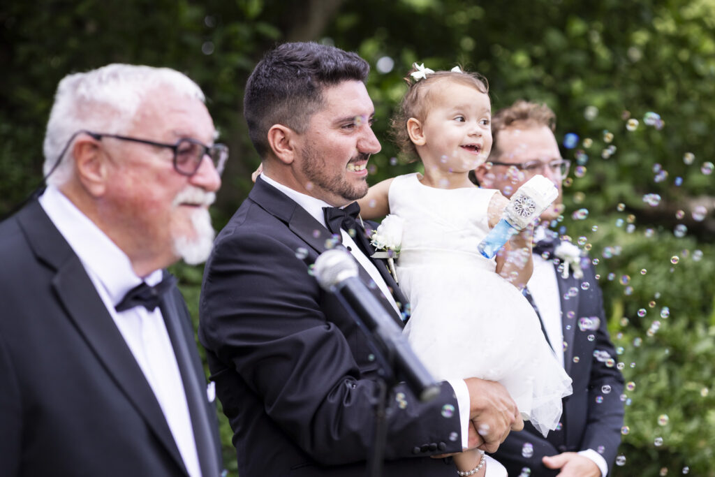 Groom holds his smiling two-year-old daughter as she plays with a bubble gun surrounded by floating bubbles during the outdoor Tyler Arboretum wedding ceremony.
