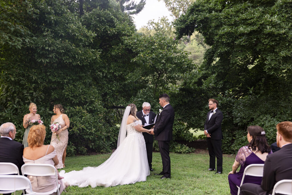 bride and groom stand hand in hand at their wedding ceremony in a lush green setting at their Tyler Arboretum wedding.