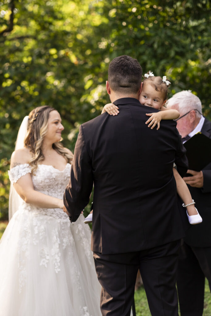 A toddler in a white dress wraps their arms around the groom’s shoulders as he carries them during the outdoor ceremony, with the bride and officiant softly out of focus against the leafy green backdrop of a Pennsylvania garden wedding.